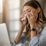 Personas en una oficina experimentando síntomas como dolores de cabeza, fatiga e irritación ocular debido al Síndrome del Edificio Enfermo.